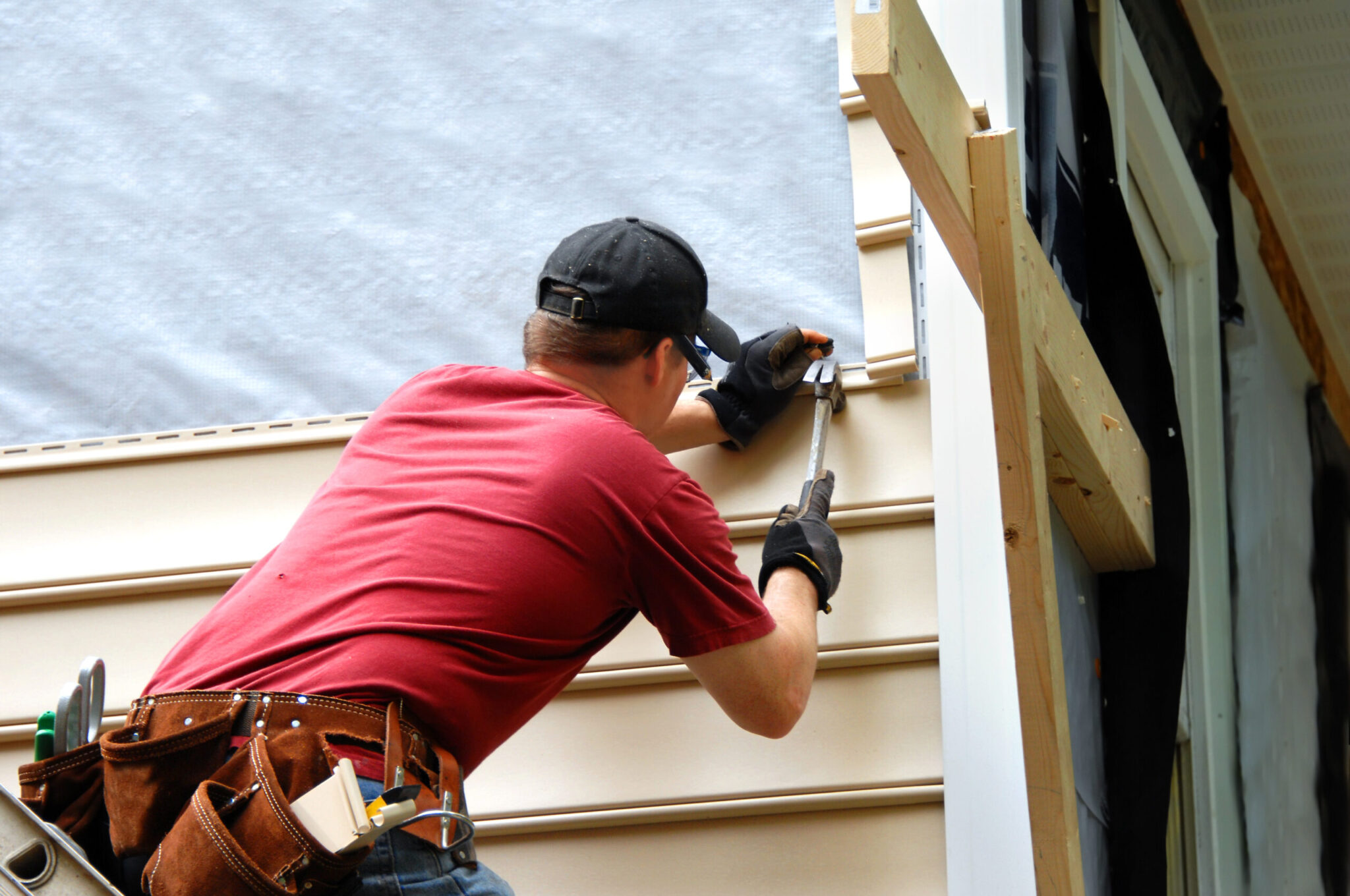 Young homeowner installs siding to his home.  He is holding a hammer and wearing a tool belt.