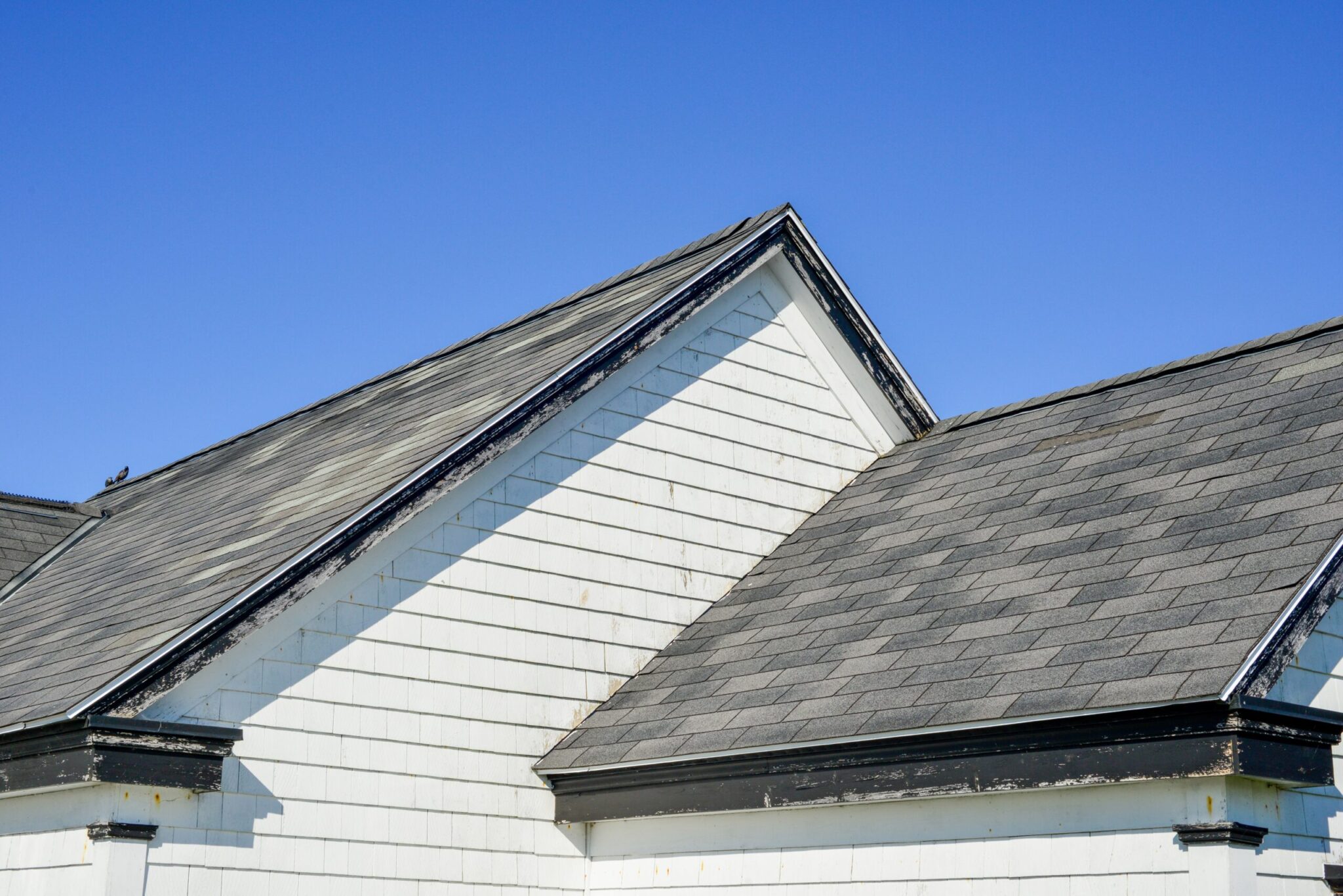 Multiple peaked and layered roofs on a white vintage wooden house. The eave is painted black. The shingles on the roof are grey in color. The old building has wood horizontal clapboard siding.